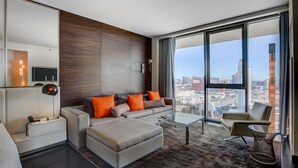Habitación de hotel moderna con un amplio ventanal que ofrece vistas a la ciudad, un sofá con cojines naranjas, una mesa de centro de cristal y una mesita de noche con lámpara.