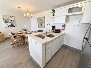 Una cocina moderna con una isla central, muebles blancos y una zona de comedor con sillas de mimbre.