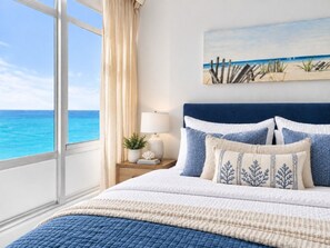 A bedroom with a large window, a bed with blue and white bedding, a nightstand with a lamp, and a painting of a beach scene.
