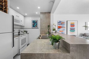 A modern kitchen with white cabinetry, a stainless steel sink, and a concrete countertop. There are two potted plants on the counter.