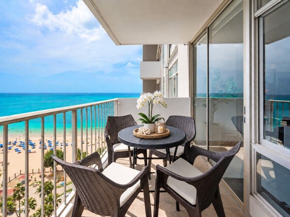 A balcony with wicker furniture, a small table with a plant, and a view of the beach and ocean.