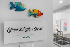 A sign reading "Sand & Wine Condo" with a "Welcome" message, two decorative fish, and a kitchen area with a sink.