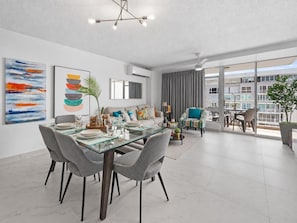 A modern living room with a glass dining table, grey upholstered chairs, a sofa, and a large window offering a view of the city.