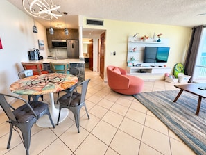 A modern living room with a round table, chairs, a pink sofa, a TV, and a dining area with a kitchen in the background.