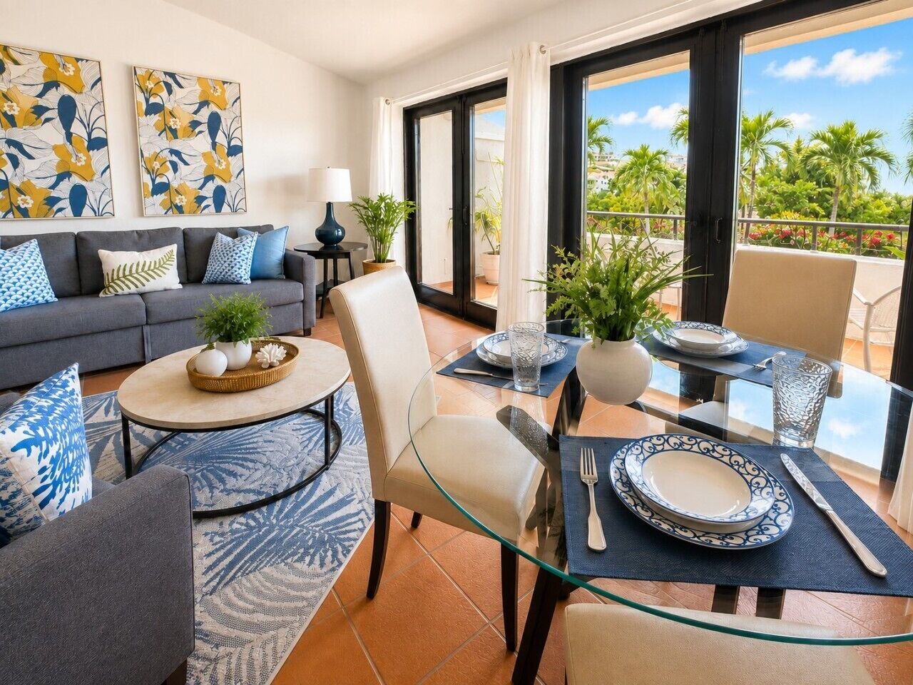 A modern living room with a glass dining table, a sofa set, and a balcony with a view of palm trees.