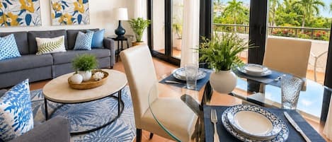 A modern living room with a glass dining table, a sofa set, and a balcony with a view of palm trees.