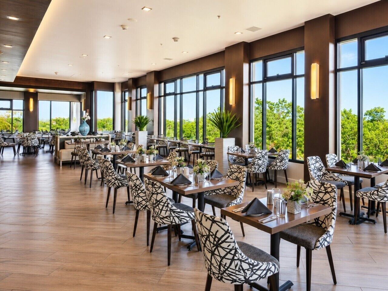 A spacious dining area with multiple tables and chairs, large windows, and a view of greenery outside.