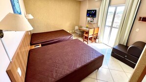 Quarto de hotel com duas camas, uma mesa com cadeira, televisão e janela com cortinas.