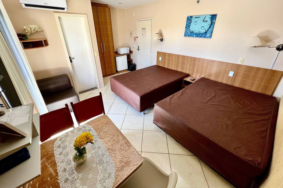 A small, well-lit room with two single beds, a desk with a chair, a small table with a vase of flowers, and a wall-mounted clock.