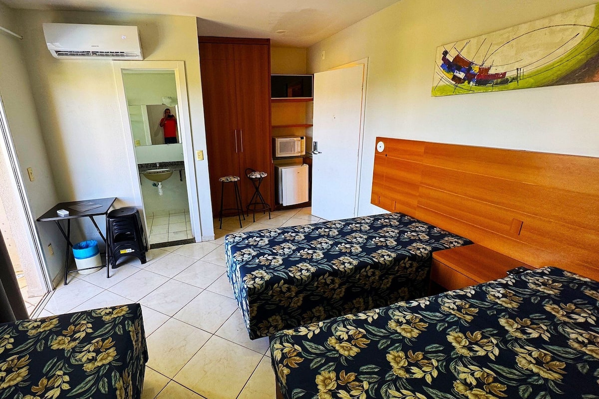 A hotel room with two beds, a small kitchenette, a bathroom, and a view of the outside.