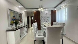 Un comedor moderno con mesa de vidrio, sillas blancas tapizadas, pared de acento de mármol y una lámpara colgante.