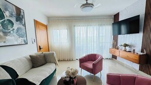 Una sala de estar moderna con un sofá verde, un sillón rosa y una mesa de centro con un jarrón de flores. Un televisor de pantalla plana está montado en la pared, y hay un mueble de madera debajo.
