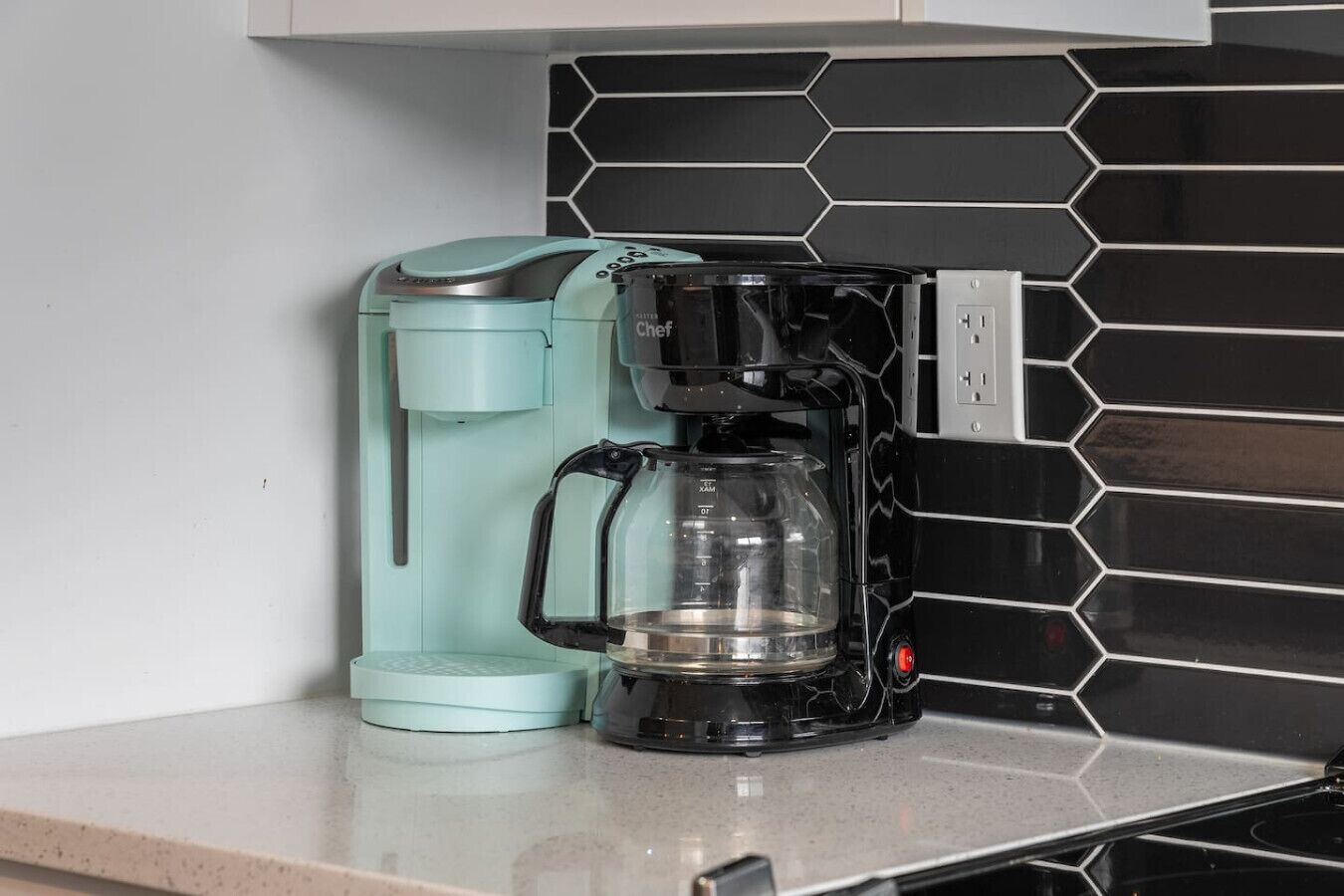 Une machine à café dotée d’une verseuse en verre transparent et d’une base de couleur turquoise, posée sur un plan de travail dont le dos est recouvert de carreaux noirs et blancs.