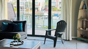 A modern living room with a sofa, a coffee table, and a chair. There is a large window with a view of buildings and a balcony outside.