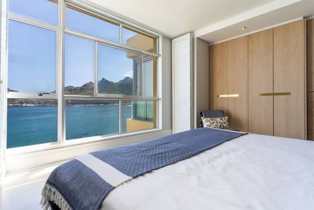 A bedroom with a large window overlooking a body of water and mountains, a bed with a blue and white striped blanket, and wooden wardrobe with gold handles.