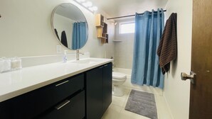 Un baño con un lavamanos blanco, armarios oscuros, un espejo redondo, un inodoro, una ducha con cortinas azules y una pequeña alfombra.