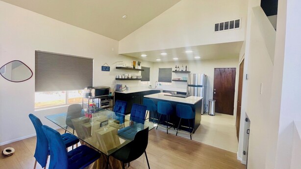 Una cocina moderna con una isla central, electrodomésticos integrados y un comedor con mesa de vidrio y sillas azules.