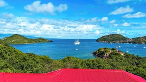 Un paisaje costero con un edificio de techo rojo, cielo azul despejado y vista al mar con barcos.