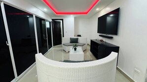 A modern living room with a white curved sofa, a flat-screen TV mounted on the wall, and a sliding glass door leading to a balcony.