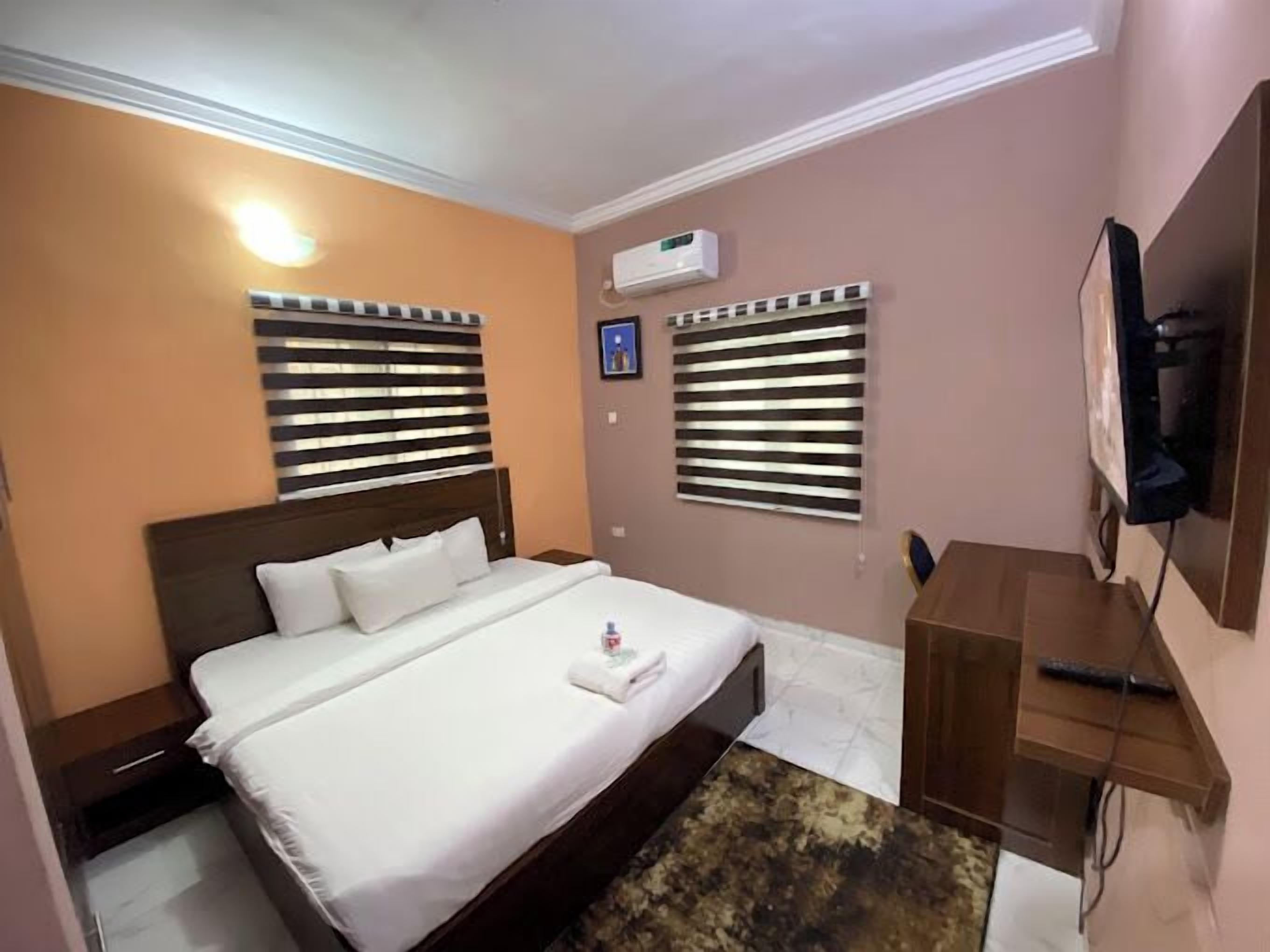 A hotel room with a bed, two windows with blinds, an air conditioning unit, a TV mounted on the wall, and a small wooden side table.