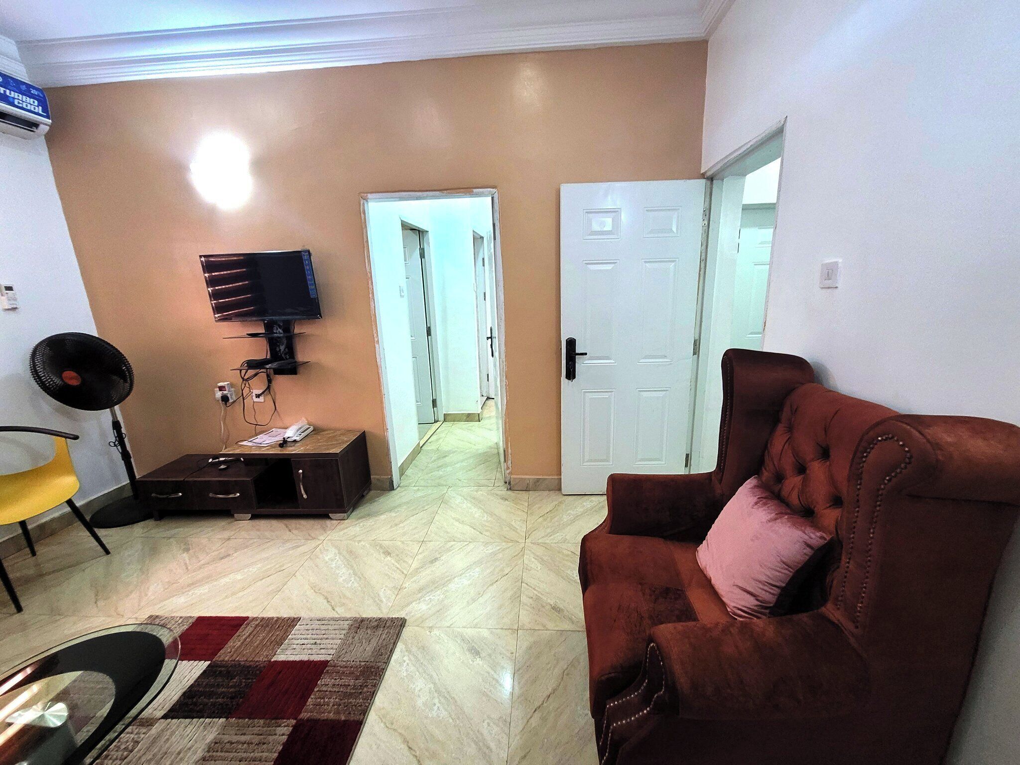 Un salon avec un canapé en cuir marron, un téléviseur fixé au mur, un ventilateur et un tapis à motifs.