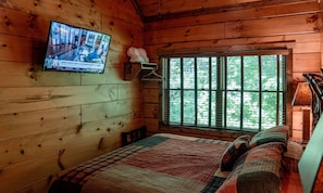 Une pièce de style cabane en bois, avec un téléviseur fixé au mur, une fenêtre donnant sur les arbres, un lit recouvert d’une courtepointe et une petite étagère ornée d’un objet décoratif.