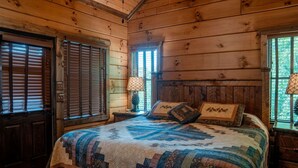 A log cabin bedroom with a bed, two windows with shutters, and a nightstand with a lamp.