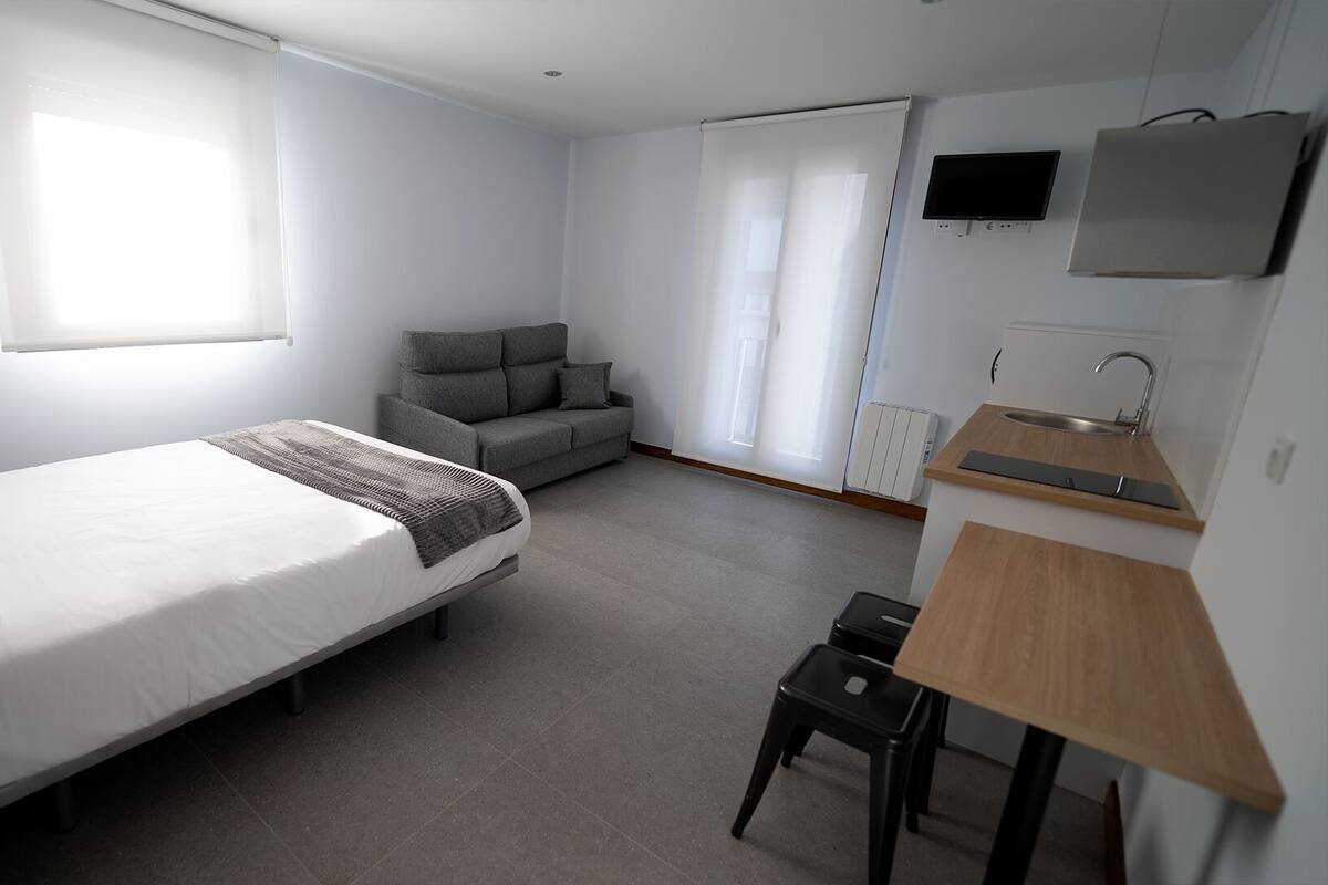 Un dormitorio moderno y bien iluminado con una cama grande, un sofá gris, un escritorio pequeño con un lavamanos y un televisor montado en la pared.