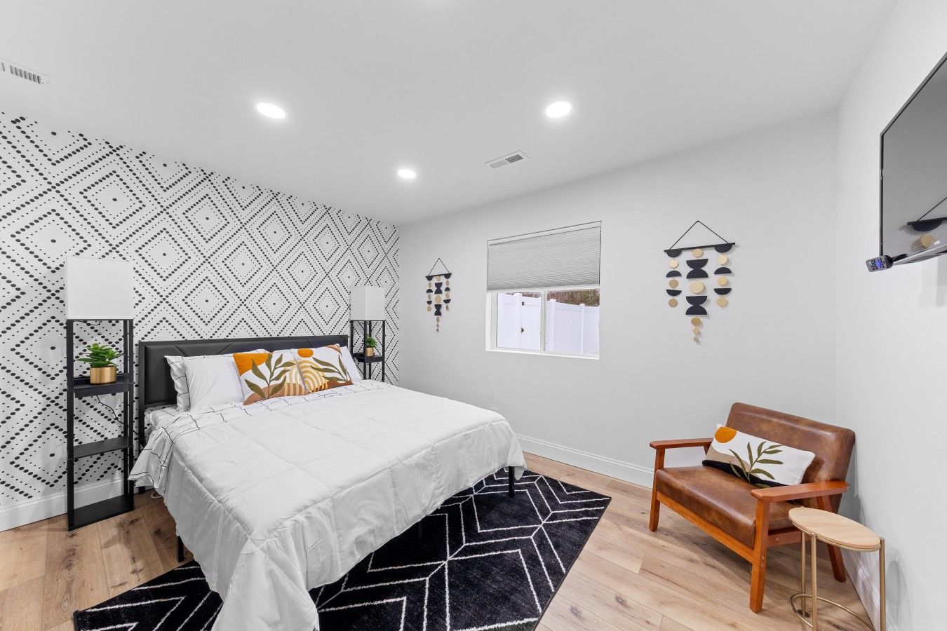 A modern bedroom with a bed, a bedside table, a chair, a window, and a wall with geometric patterns.