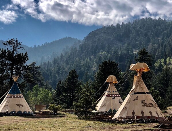 Three traditional tipis stand in a serene natural setting, surrounded by dense forested hills.