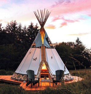Relax in a traditional teepee-style tent with a cozy fire pit and Adirondack chairs on a wooden deck.