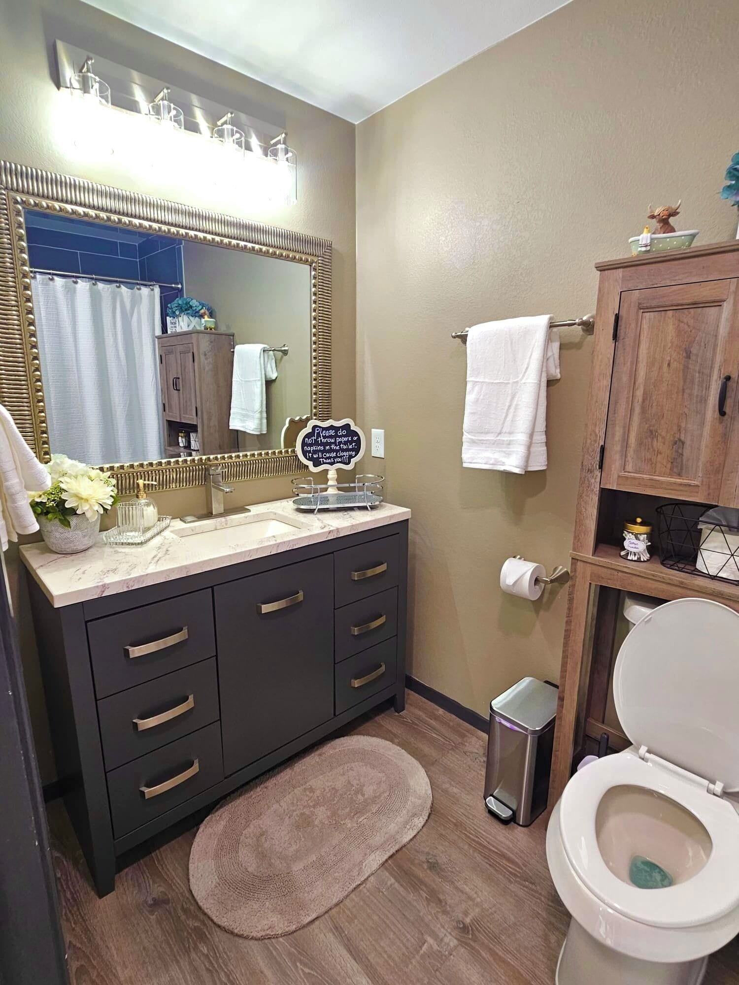 A bathroom with a vanity, mirror, sink, and toilet. There is a sign on the mirror, a towel rack, and a trash can.