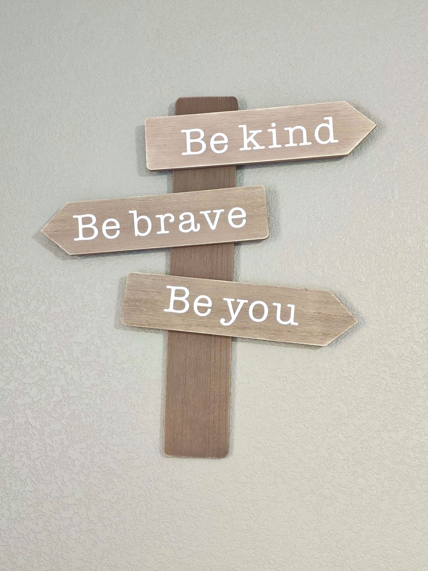 A wooden sign with three arrows pointing in different directions, each with a motivational phrase: "Be kind," "Be brave," and "Be you."