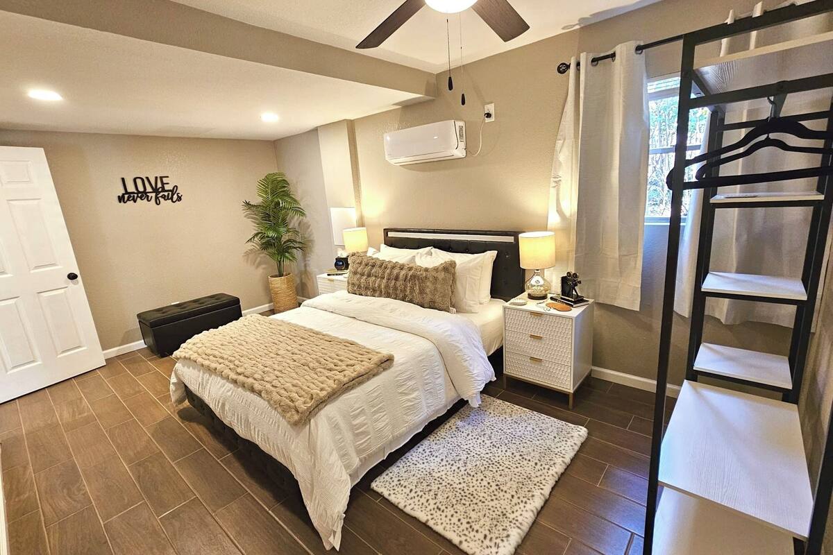 Un dormitorio con cama, mesitas de noche, ventilador de techo, equipo de aire acondicionado, planta en maceta y ventana con cortinas.