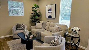 Een woonkamer met een beige bank, een zwarte salontafel en een fauteuil met een patroon. Er staat een grote plant, een kunstwerk in een lijst en een vaas met bloemen op een bijzettafeltje.