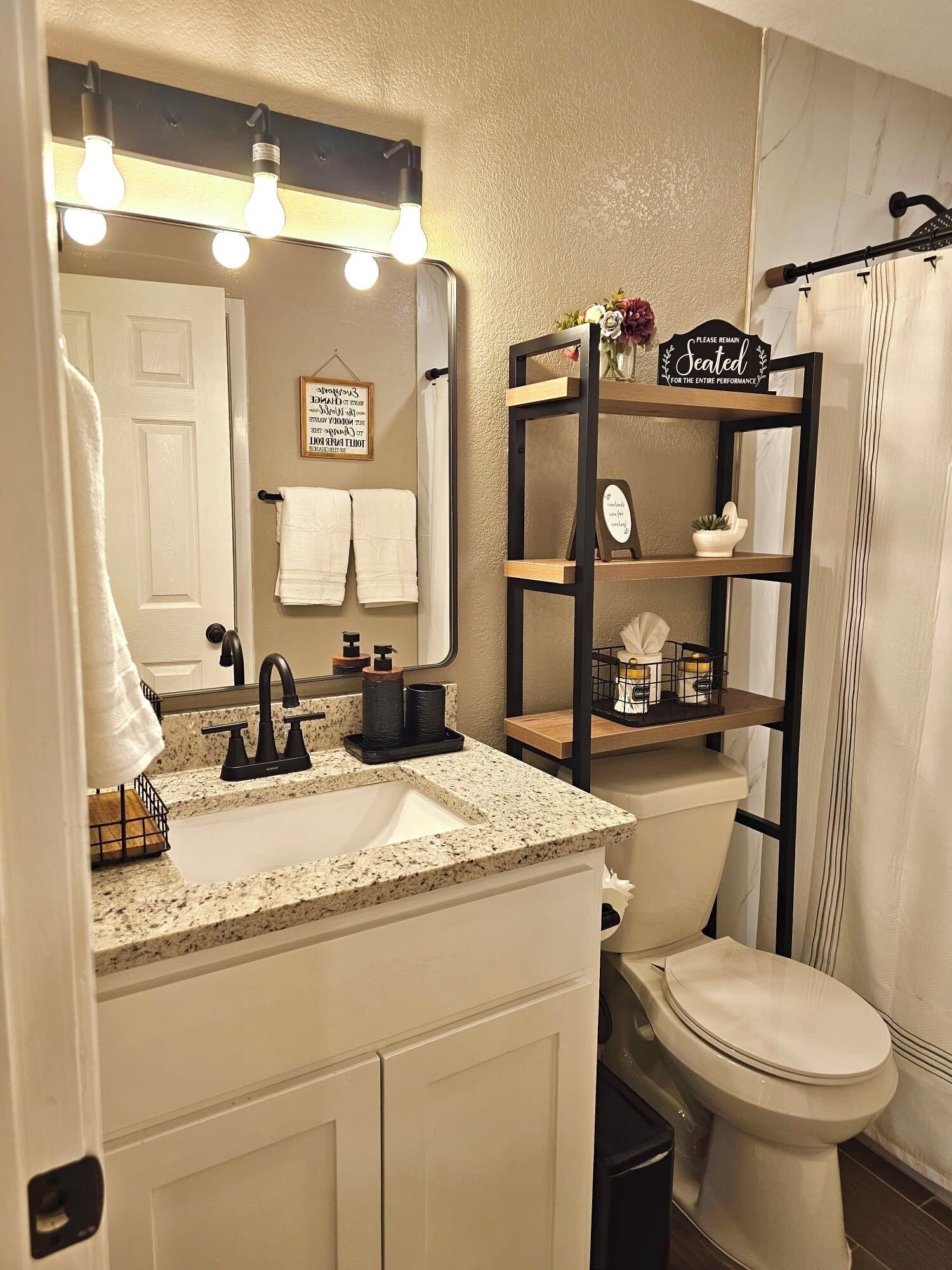 A bathroom with a sink, mirror, and toilet. There is a towel rack with two towels, a shelf with toiletries, and a sign that says "Please remain seated for the entire performance".