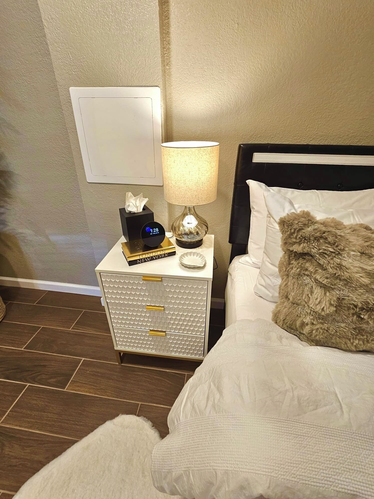 Ein ordentlich eingerichtetes Hotelzimmer mit einem Bett, einem Nachttisch mit Lampe, einer Uhr und einer Taschentuchbox.