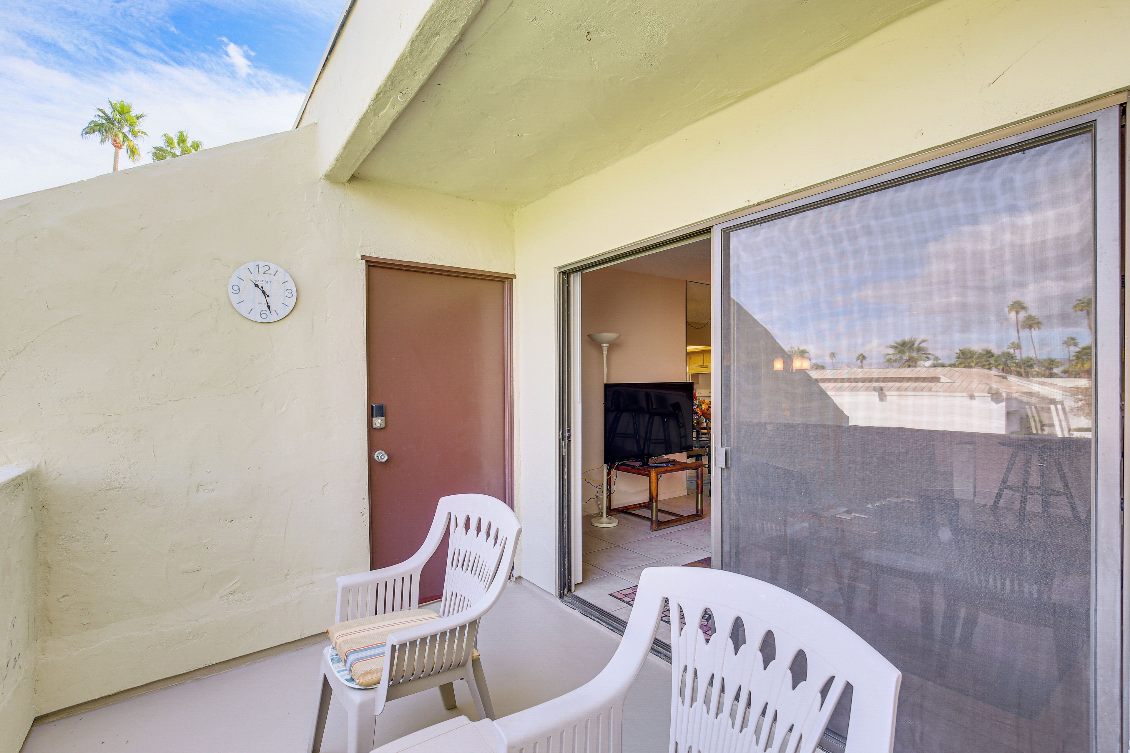 A balcony with white plastic chairs, a clock, and a sliding glass door leading to a room with a TV and a table.