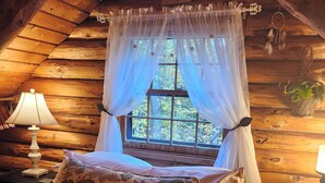 A cozy bedroom with a wooden ceiling, a canopy bed with floral pillows, a bedside lamp, and a window with a view of trees. - Lovely Log Cabin Retreat: Cozy Fireplace, Beautiful Gardens & Nature (Bradford)