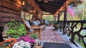 A wooden cabin with a porch featuring wicker furniture, potted plants, and an American flag. - Lovely Log Cabin Retreat: Cozy Fireplace, Beautiful Gardens & Nature (Bradford)