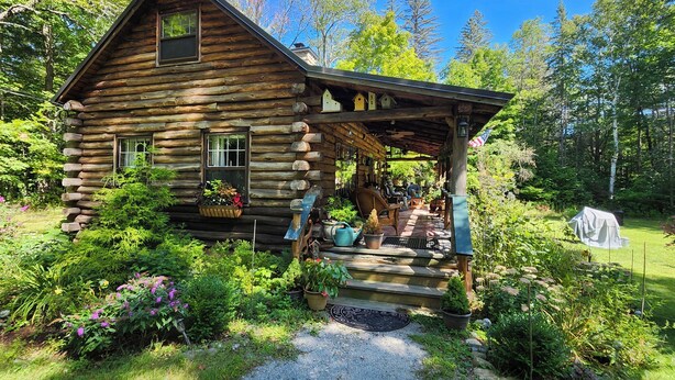 A log cabin with a porch, surrounded by lush greenery and trees. - Lovely Log Cabin Retreat: Cozy Fireplace, Beautiful Gardens & Nature (Bradford)