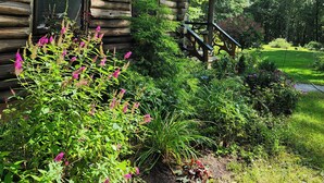 A log cabin with a porch, surrounded by lush greenery and flowering plants. - Lovely Log Cabin Retreat: Cozy Fireplace, Beautiful Gardens & Nature (Bradford)