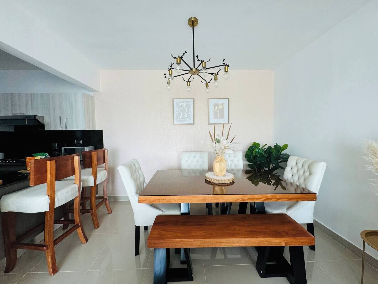 A dining area with a wooden table, white upholstered chairs, a bench, a chandelier, and a potted plant.