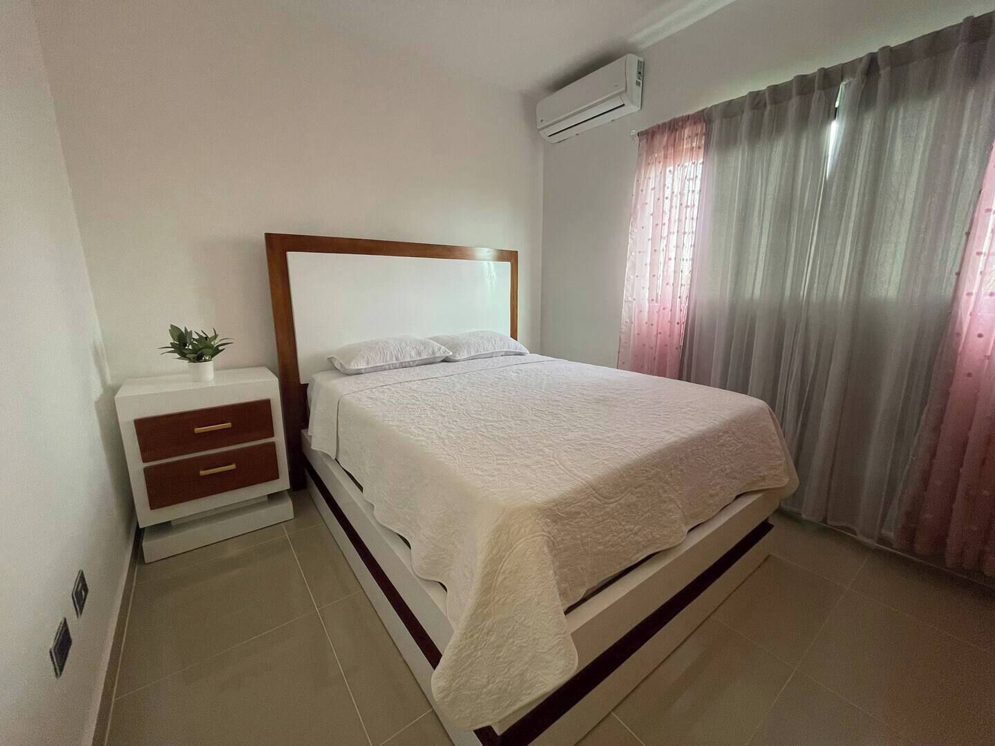 A neatly arranged bedroom with a bed, bedside tables, a small plant, and an air conditioning unit.