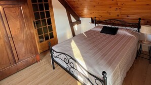 Quarto com teto de madeira, cama com estrutura metálica, guarda-roupa de madeira e janela com grade.