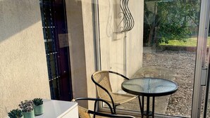 Uma área de pátio com porta de vidro, armário branco com gavetas, cadeiras de vime, mesa redonda e um abajur de metal decorativo.