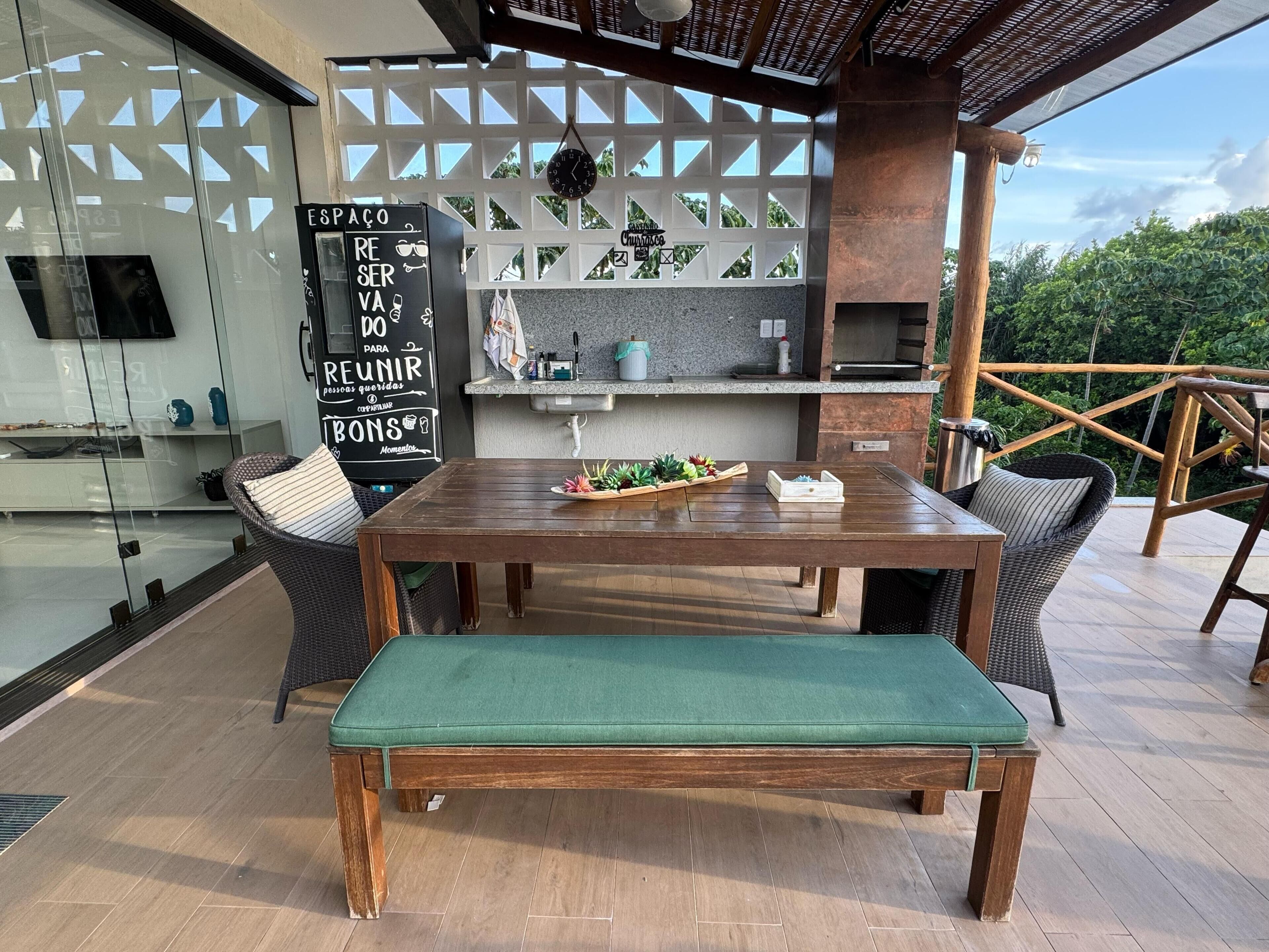 An outdoor dining area with a wooden table and bench, a kitchen area with a grill, and a view of greenery.