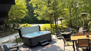 Un jacuzzi en una terraza de madera con mobiliario exterior y vistas a una zona boscosa.