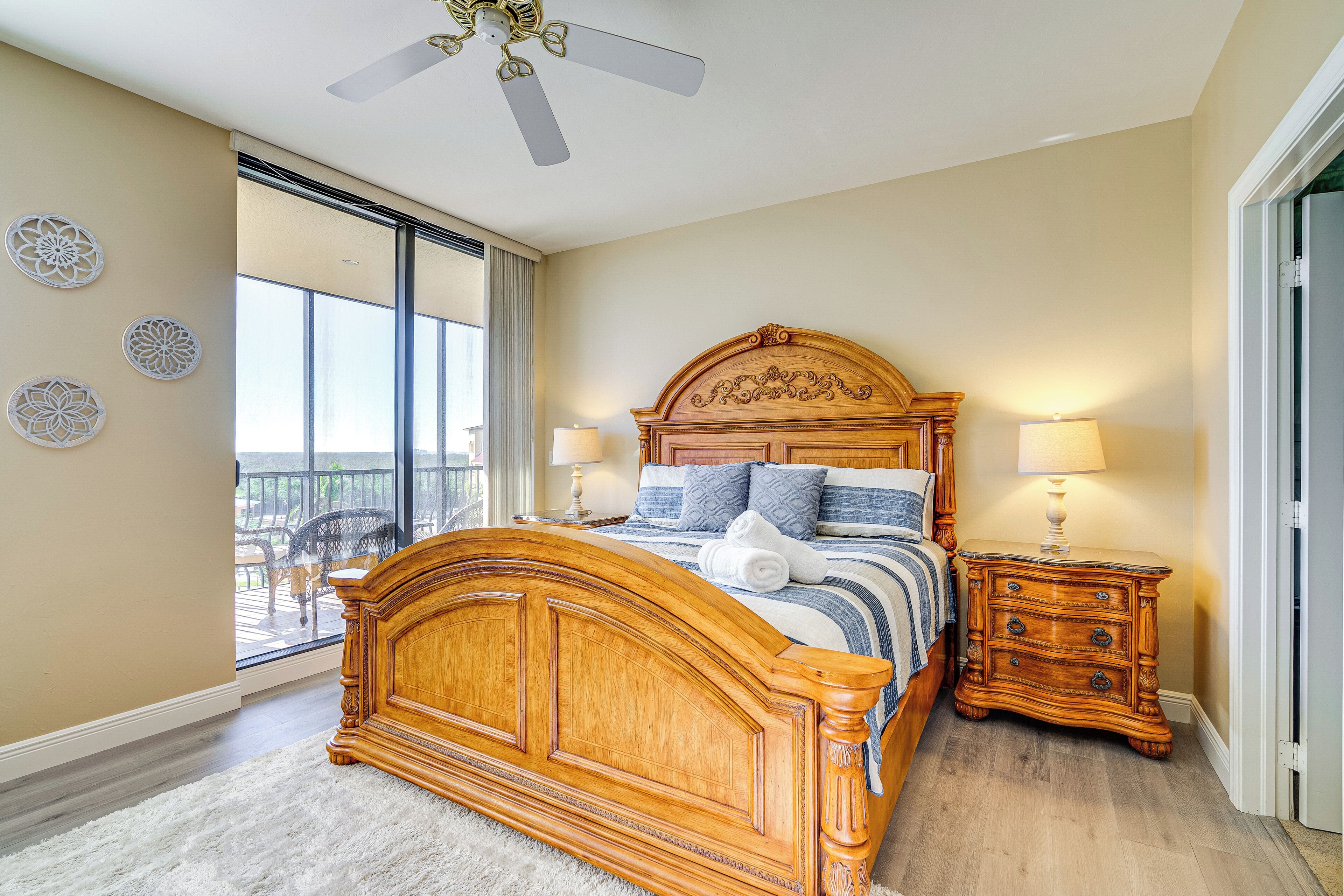 A bedroom with a wooden bed, two bedside tables with lamps, a ceiling fan, and a balcony with outdoor furniture.
