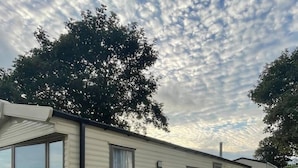 Una casa móvil con fachada blanca, un porche cercado y un árbol grande al fondo.